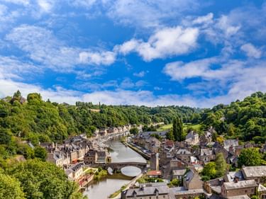 Luftaufnahme von Dinan mit Steinbrücke über den Fluss, historischen Gebäuden mit grauen Dächern am Ufer, umgeben von grünen Hügeln.