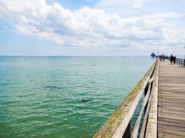 Hölzerne Seebrücke, die sich in die türkisfarbene Ostsee in Grömitz erstreckt. Menschen spazieren auf der Brücke unter bewölktem Himmel.