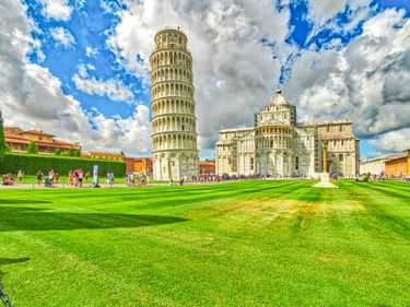 Der Schiefe Turm von Pisa und der Dom von Pisa auf grünem Rasen unter blauem Himmel mit weißen Wolken. Touristen auf dem Rasen sichtbar.