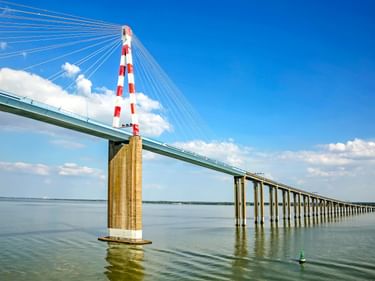 Saint-Nazaire-Brücke mit rot-weiß gestreiftem Pylon und Schrägseilen über der Loire-Mündung unter blauem Himmel mit weißen Wolken.