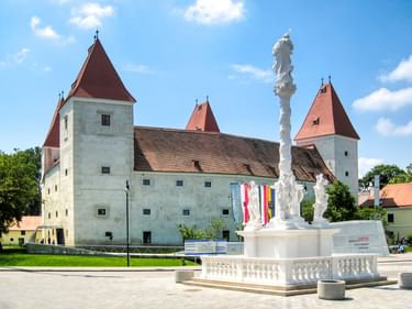Schloss Orth mit roten Kegeldächern und weißen Mauern, mit einer hohen weißen barocken Säulenstatue im Vordergrund.