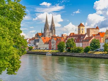 View of Regensburg's historic old town with twin-spired cathedral and medieval stone bridge tower along the Danube River under blue sky.