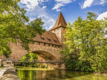 Historischer Kettensteg mit Steinturm und Bogen über einem Fluss in Nürnberg, umgeben von grünen Bäumen unter blauem Himmel.