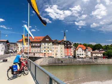 Zwei Radfahrer auf einer Brücke über die Isar in Bad Tölz, mit bunten historischen Gebäuden und einem Kirchturm am gegenüberliegenden Ufer.