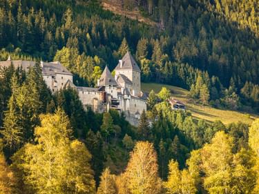 Schloss Moosham mit Türmen und grauen Mauern auf einem Hang, umgeben von dichtem Wald mit goldener Herbstfärbung in Österreich.