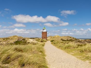 Roter Backsteinleuchtturm Quermarkenfeuer mit grüner Laterne auf Sanddünen. Ein Kiesweg führt durch Dünengras zum Turm unter blauem Himmel.