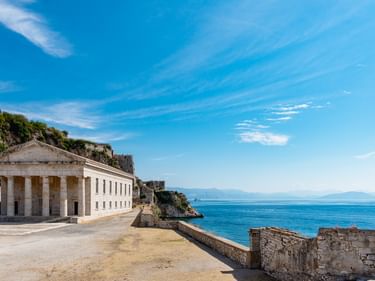 Kirche des Agios Georgios mit neoklassizistischen Säulen an Korfus Küste. Blaues Meer und Himmel sichtbar, Ruinen im Vordergrund, Klippen dahinter.