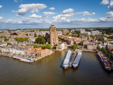 Luftaufnahme von Dordrecht mit hohem Kirchturm, historischen Gebäuden mit orangefarbenen Dächern und Frachtschiffen am Wasserweg unter blauem Himmel.