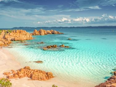 Sandstrand mit türkisfarbenem Wasser und rötlichen Felsformationen in der Bucht La Maddalena. Klares flaches Wasser zeigt Sandmuster.