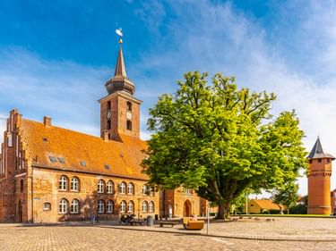 Backsteinkirche mit orangefarbenem Ziegeldach und Turm in Nykøbing Falster. Großer grüner Baum auf Kopfsteinpflasterplatz mit Wasserturm.