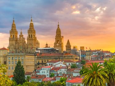 Die Türme der Kathedrale von Santiago de Compostela erheben sich bei Sonnenuntergang über Gebäude mit roten Dächern. Palmen rahmen die Altstadt.