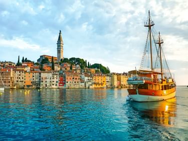Traditionelles Holzboot im Hafen von Rovinj mit bunten historischen Gebäuden und Kirchturm auf dem Hügel unter bewölktem Himmel.