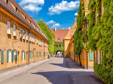 Schmale Straße in der Augsburger Fuggerei mit gelben Gebäuden mit grünen Fensterläden, efeubedeckten Wänden und Kirchturm unter blauem Himmel.