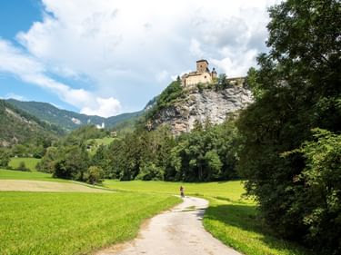 Radfahrer auf Weg durch grüne Wiesen zum Schloss Ortenstein auf Felsenklippe, umgeben von bewaldeten Bergen unter blauem Himmel.