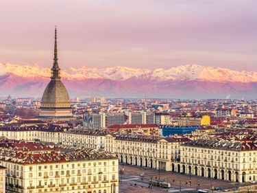Panoramablick auf Turin mit dem Turm der Mole Antonelliana, der Piazza Vittorio Veneto und schneebedeckten Alpen im Hintergrund unter rosa Abendhimmel.
