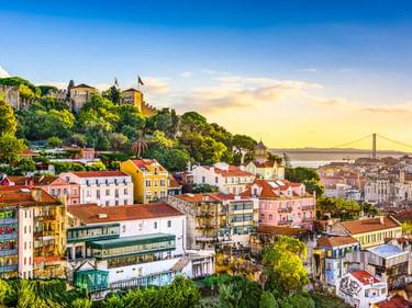 Bunte Häuser am Hang von Lissabon mit der Burg Castelo de São Jorge oben. Eine Hängebrücke überspannt den Fluss im Hintergrund bei Sonnenuntergang.
