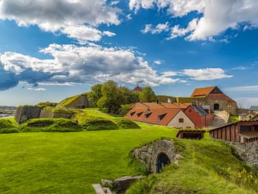 Festung Varberg mit grasbewachsenen Wällen, steinernem Tunneleingang und historischen Gebäuden mit roten Ziegeldächern unter blauem Himmel.
