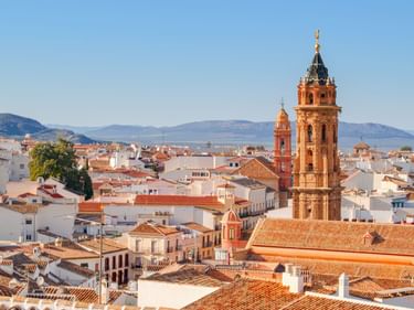 Panoramablick auf Antequera mit weißen Gebäuden und Terrakottadächern. Ein markanter Kirchturm ragt über die Stadtlandschaft mit Bergen im Hintergrund.