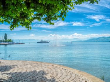 Gepflasterte Promenade am Bodensee in Arbon mit Fährschiff auf ruhigem blauen Wasser, Baumzweigen darüber und fernen Bergen unter blauem Himmel.