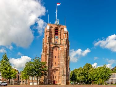 Der Oldehove, ein hoher schiefer Backsteinturm in Leeuwarden mit niederländischer Flagge, umgeben von Bäumen und Gebäuden unter blauem Himmel.