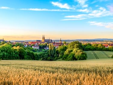 Panoramablick auf Bamberg mit goldenem Weizenfeld im Vordergrund, historischer Altstadt mit Kirchtürmen und grünen Feldern unter blauem Himmel.
