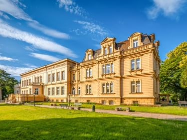 Gelbes neoklassizistisches Schloss von Ostromecko mit verzierter Fassade, umgeben von grünem Rasen und Bäumen unter blauem Himmel.