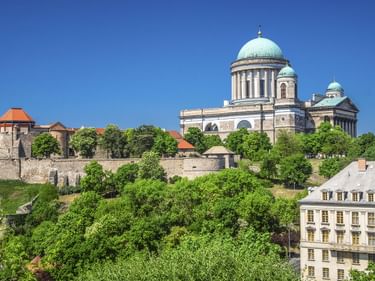 Die Basilika von Esztergom mit grünen Kupferkuppeln erhebt sich über Festungsmauern und üppigen Bäumen. Ein historisches Gebäude mit Mansardendach im Vordergrund.