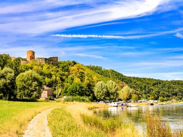 Radweg entlang der Weser zwischen Polle und Bodenwerder mit Burgruine auf bewaldetem Hügel, Boote am Flussufer vertäut.
