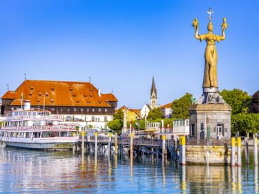 Imperia-Statue am Konstanzer Hafen mit Konzilgebäude, Kirchturm und Fahrgastschiff. Klarer blauer Himmel und ruhiges Wasser mit Holzpfählen.