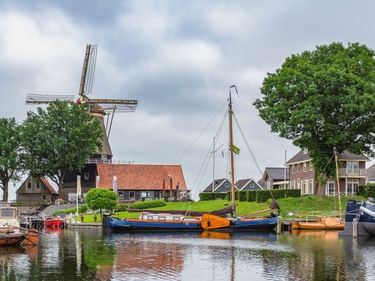 Holländische Windmühle mit rotem Ziegeldach neben einem Kanal in Harderwijk. Traditionelle Boote am Ufer mit Häusern und Bäumen.