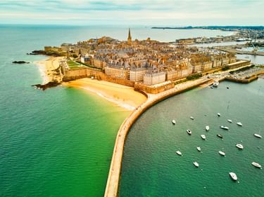 Luftaufnahme der ummauerten Altstadt von Saint-Malo auf einer Halbinsel, umgeben von türkisfarbenem Meer mit Booten im Hafen und Sandstränden.
