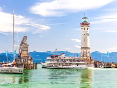 Lindauer Hafeneinfahrt am Bodensee mit weißem Leuchtturm, bayerischer Löwenstatue auf Sockel und Raddampfer. Berge im Hintergrund sichtbar.