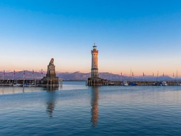Lindau harbor entrance with lighthouse and lion statue on stone pedestals in Lake Constance. Calm water reflects the structures under clear sky.