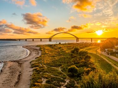 Fehmarnsundbrücke mit markanter Bogenform über dem Wasser bei Sonnenuntergang. Küstenweg und Strand im Vordergrund, goldener Himmel.