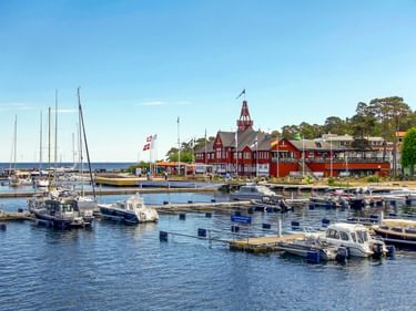 Sandhamn Hafen mit Booten an Holzstegen. Rote Holzgebäude mit Turm am Ufer unter klarem blauen Himmel.