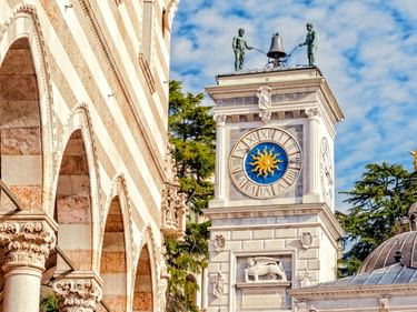 Weißer Glockenturm in Udine mit blau-goldenem Sonnenzifferblatt, gekrönt von zwei Bronzestatuen und einer Glocke. Gestreiftes Gebäude links.
