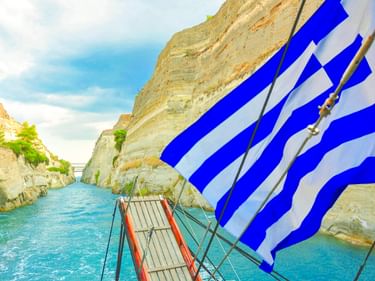 Blick vom Bootsdeck mit griechischer Flagge im Vordergrund, fahrend durch den schmalen Kanal von Korinth mit steilen Kalksteinwänden.