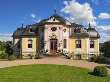 Barockschloss mit rosa und gelber Fassade, prunkvollem Eingang und symmetrischen Flügeln. Gepflegter Rasen und Kiesweg führen zum Gebäude.
