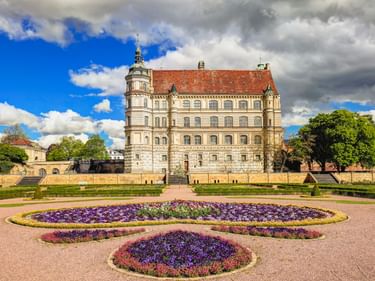 Renaissance-Schloss Güstrow mit rotem Ziegeldach und Turm, Blick über Ziergarten mit runden lila und rosa Blumenbeeten unter dramatischem Himmel.