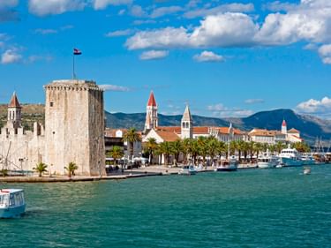 Trogir Altstadt Uferpromenade mit steinerner Festungsturm, Kirchtürmen, palmengesäumter Promenade und Booten im türkisfarbenen Hafen.