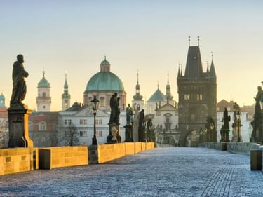 Karlsbrücke in Prag bei Sonnenaufgang mit barocken Statuen entlang des Kopfsteinpflasters. Historische Türme und Kuppelkirchen im Hintergrund.