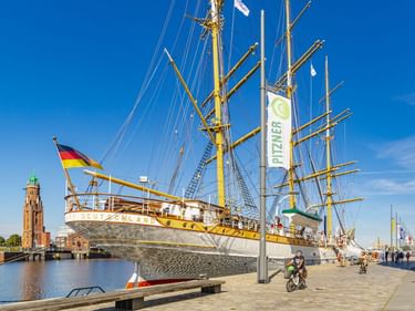 Weißes Segelschiff Deutschland im Neuen Hafen in Bremerhaven. Roter Backsteinleuchtturm links, Radfahrer auf Promenade unter blauem Himmel.