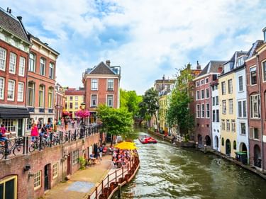 Kanal in Utrecht gesäumt von bunten historischen Gebäuden. Menschen sitzen an Ufenterrasse mit gelben Sonnenschirmen. Bäume und Fahrräder entlang des Kanals.