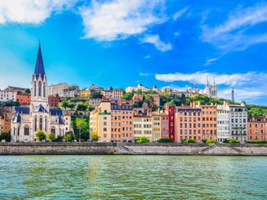 View across the Rhône River in Lyon showing colorful historic buildings and a white church with a tall spire under a blue sky.