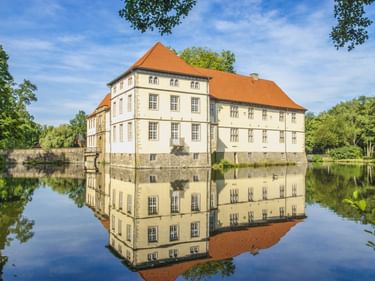 Schloss Strünkede, ein cremefarbenes Wasserschloss mit orangefarbenem Ziegeldach in Herne, perfekt im umgebenden Wasser gespiegelt.