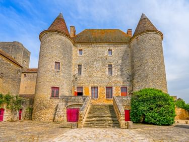 Château de Nemours in Frankreich mit zwei runden Türmen, Steintreppe, roten Türen und Kopfsteinpflasterhof unter blauem Himmel.