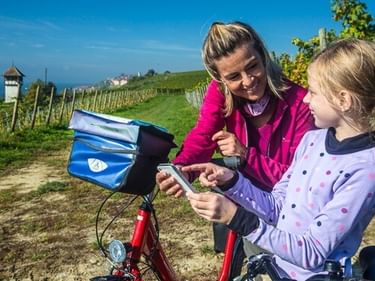 Woman and girl with bicycle looking at smartphone in vineyard near Meersburg. Lake Constance and watchtower visible in background.