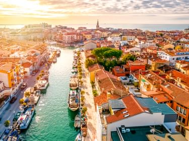 Luftaufnahme von Grado, Italien bei Sonnenuntergang mit Kanal und Booten, bunten Gebäuden mit roten Ziegeldächern und der Adria im Hintergrund.