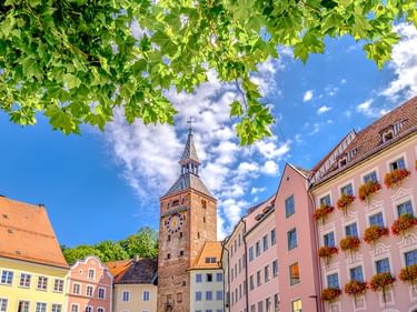 Historischer Schmalzturm in Landsberg am Lech, umgeben von bunten Gebäuden mit Blumenkästen unter grünen Baumzweigen und blauem Himmel.