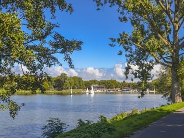 Blick auf den Aasee in Münster mit Segelbooten auf ruhigem Wasser, eingerahmt von grünen Bäumen. Ein asphaltierter Weg verläuft am Ufer.
